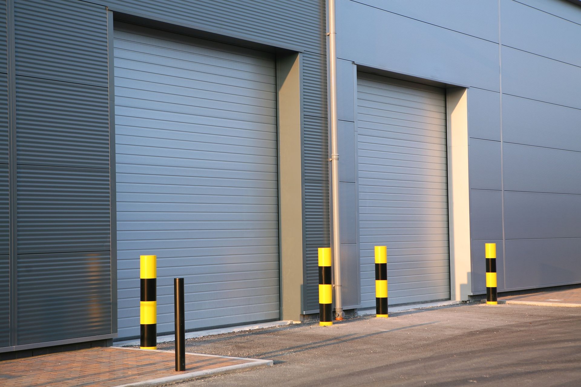 Two gray industrial roll-up doors with black and yellow bollards in front.