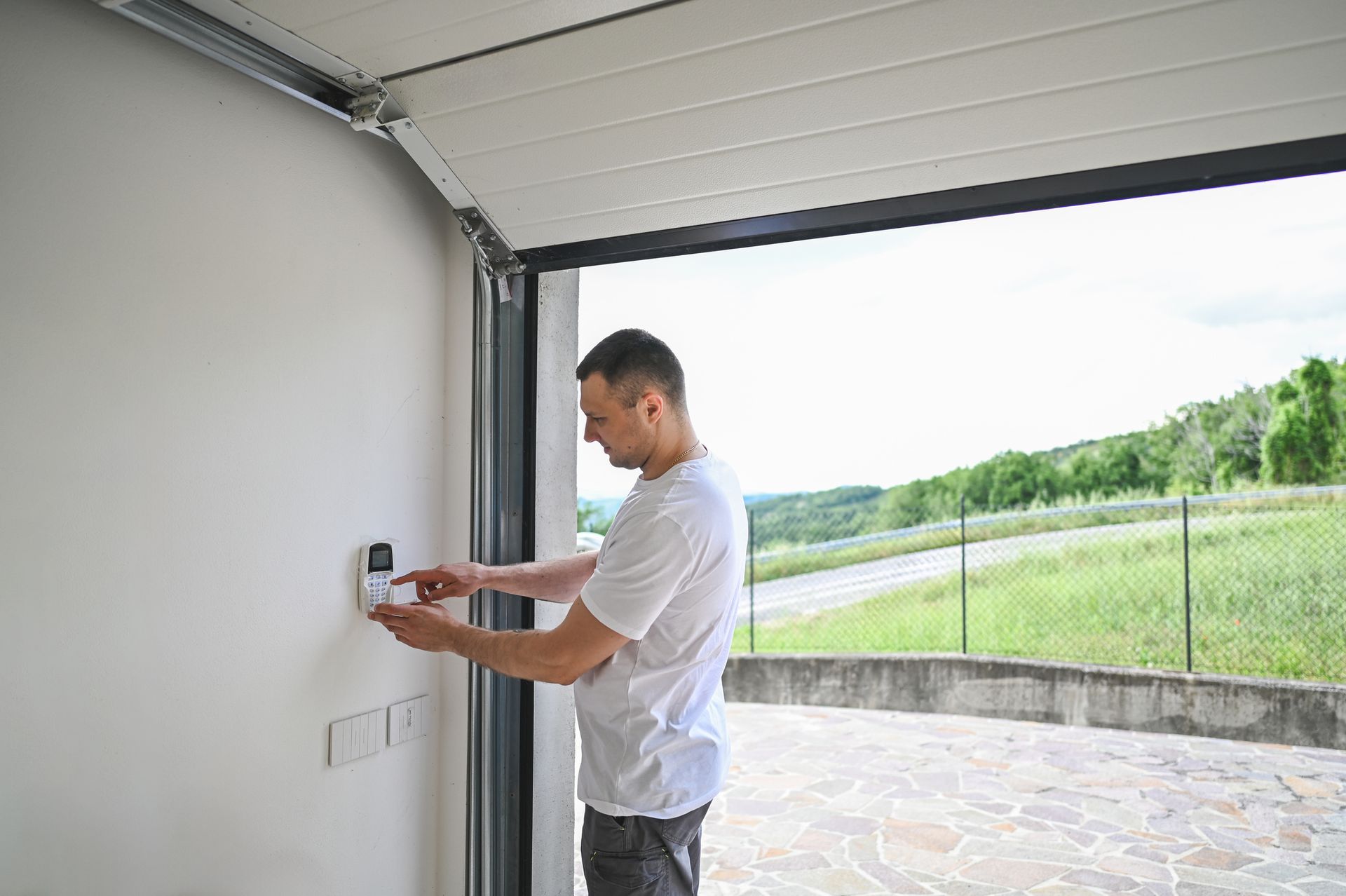 Man in garage adjusts a wall-mounted device, garage door open to scenic outdoors.