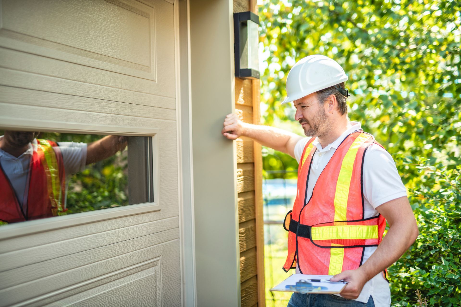 Construction worker in hard hat and vest, inspecting a garage door, holding a clipboard.