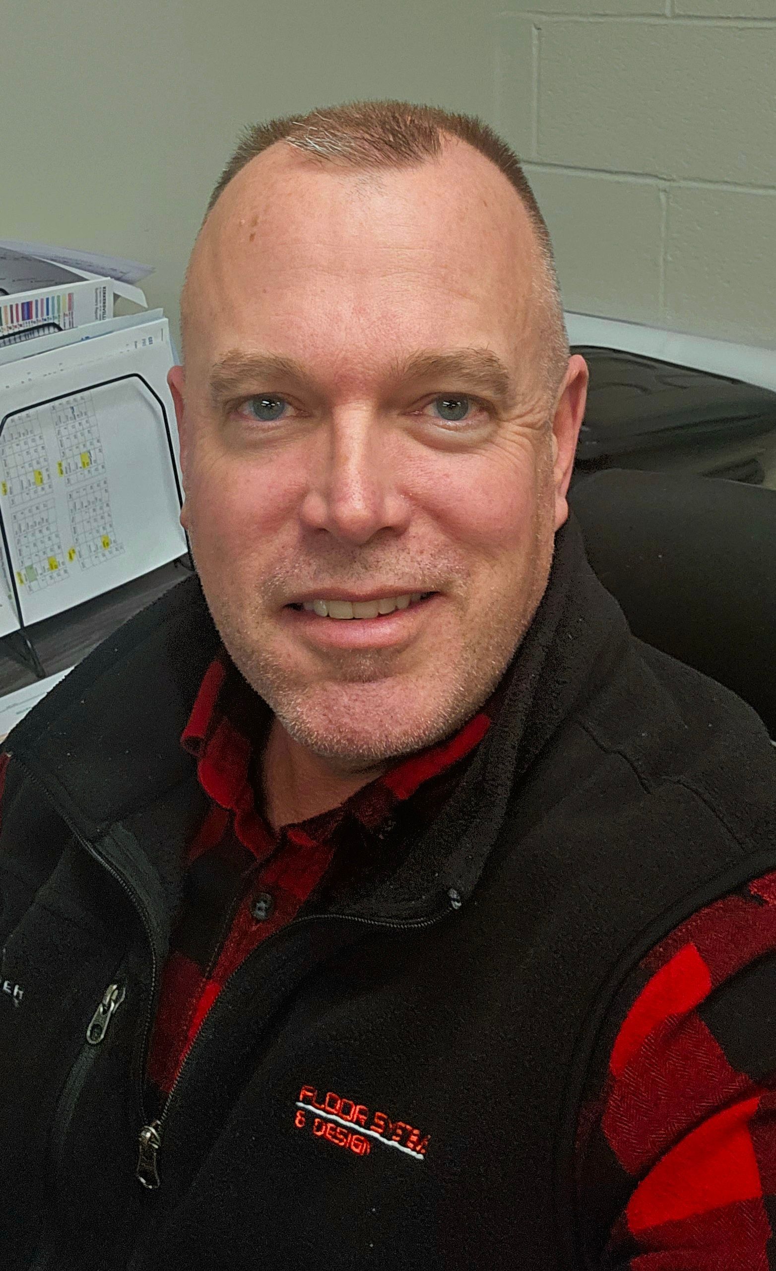 Man smiling in a black vest and red plaid shirt, seated in an office cubicle