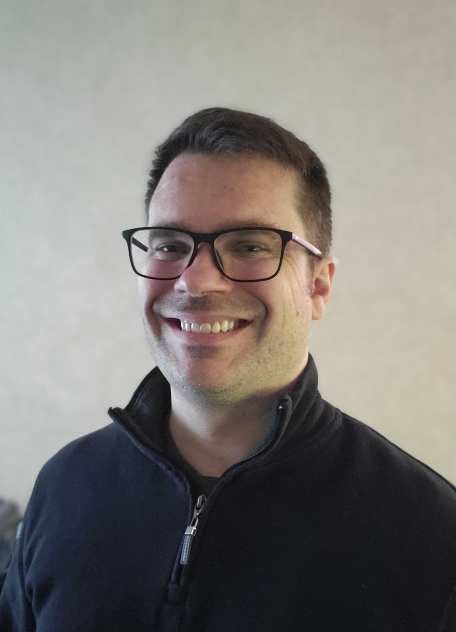 Smiling person with glasses and a dark zip-up jacket against a light gray background