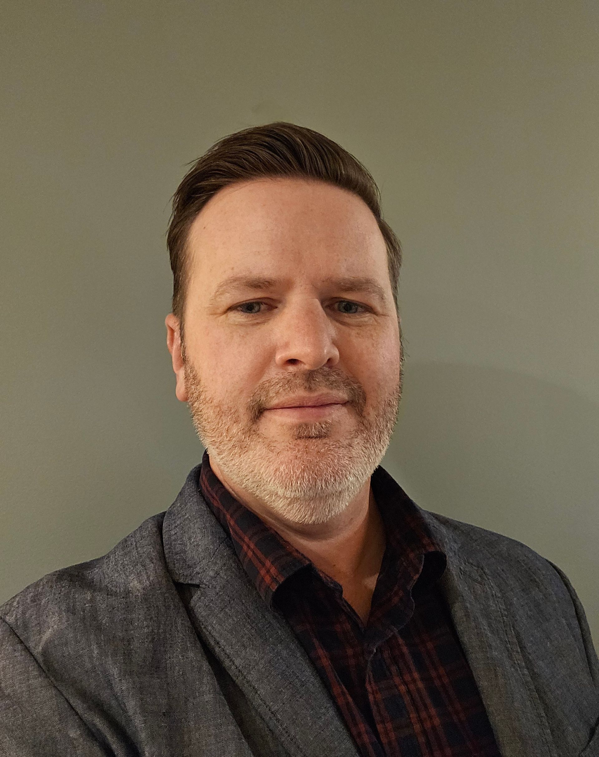 Portrait of a man in a gray blazer over a dark plaid shirt, standing against a plain wall.