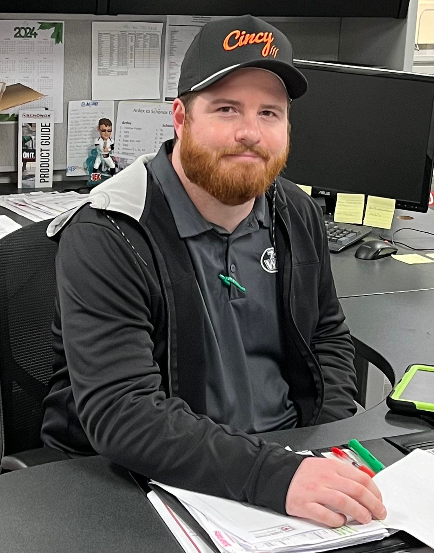 Man in black cap and shirt sits at a desk, smiling and signing a paper in an office.