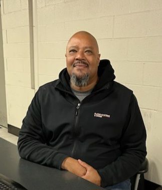 Man in a black hoodie seated at a table against a light cinderblock wall, smiling gently.