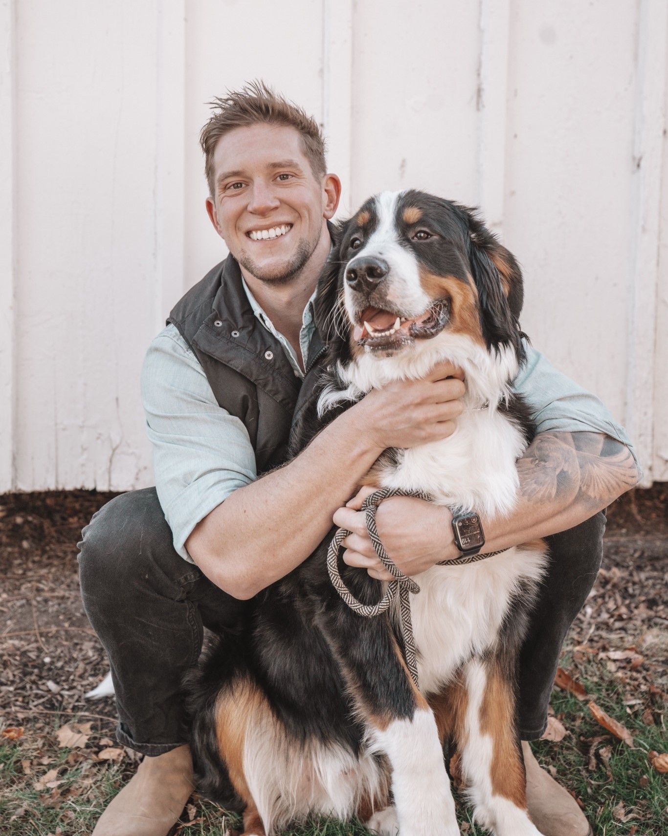 Smiling person hugging a large black, white, and brown dog outdoors in front of a white wall