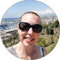 A person wearing sunglasses smiles in a selfie outdoors with a blurred coastal city and harbor in the background.