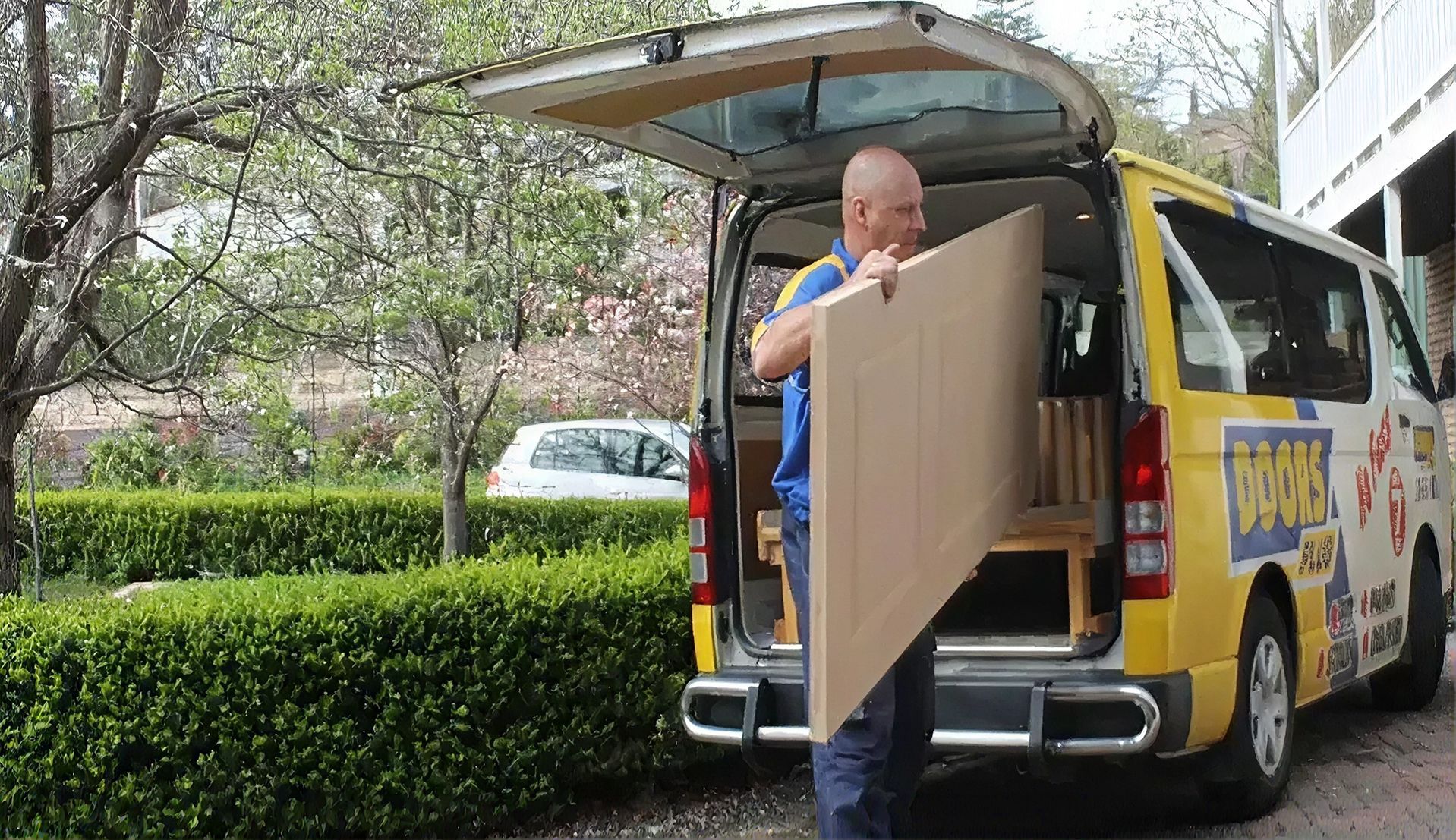 Man loading a large panel into a yellow van on a tree-lined street