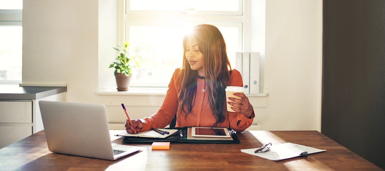 Person working at a desk with a laptop, tablet, and coffee in a bright home office