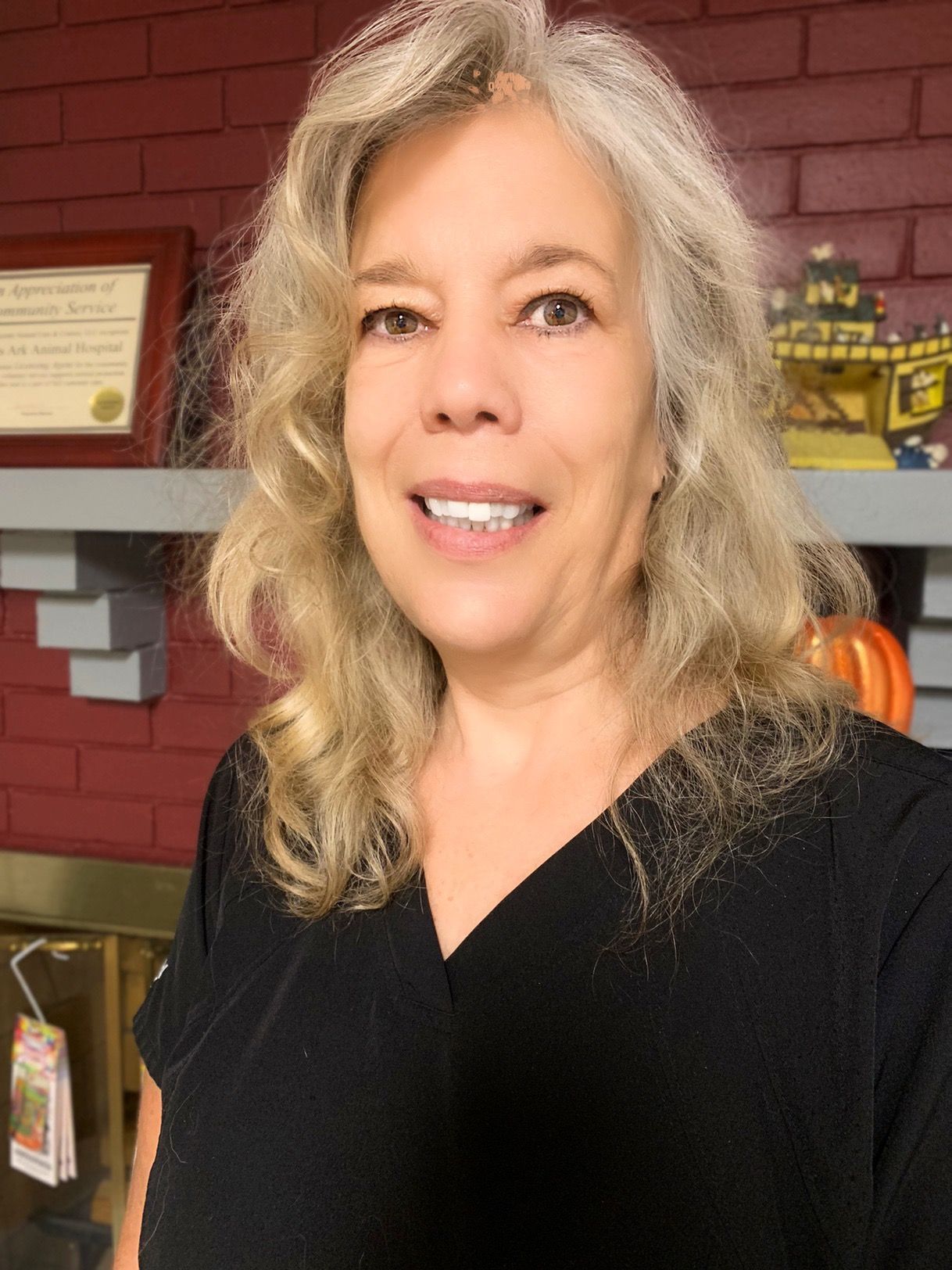 A woman in a black scrub top is smiling in front of a fireplace.