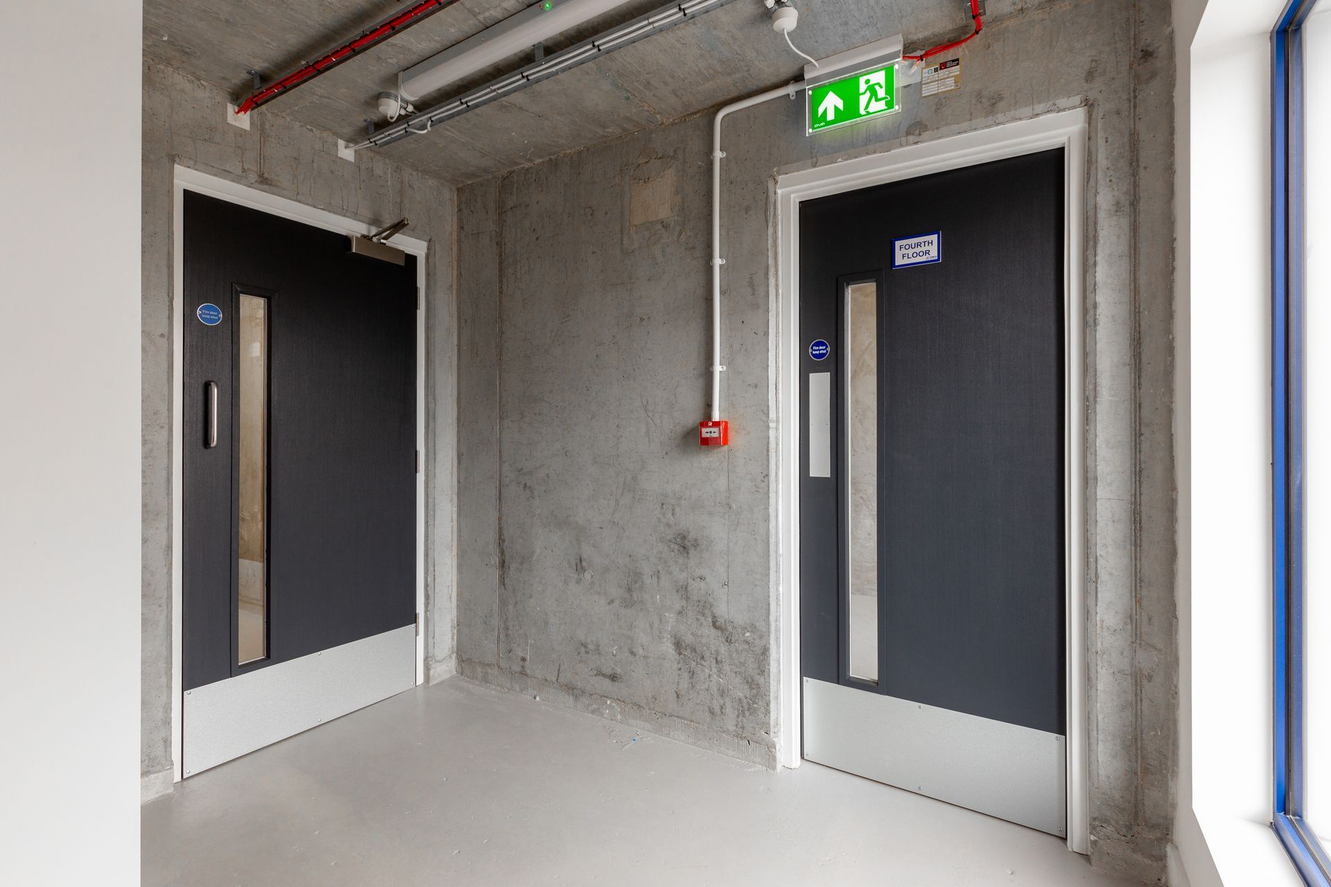 Two fire doors in a concrete hallway with an exit sign and a fire alarm button on the wall. Two fire doors in a concrete hallway with an exit sign and a fire alarm button on the wall.