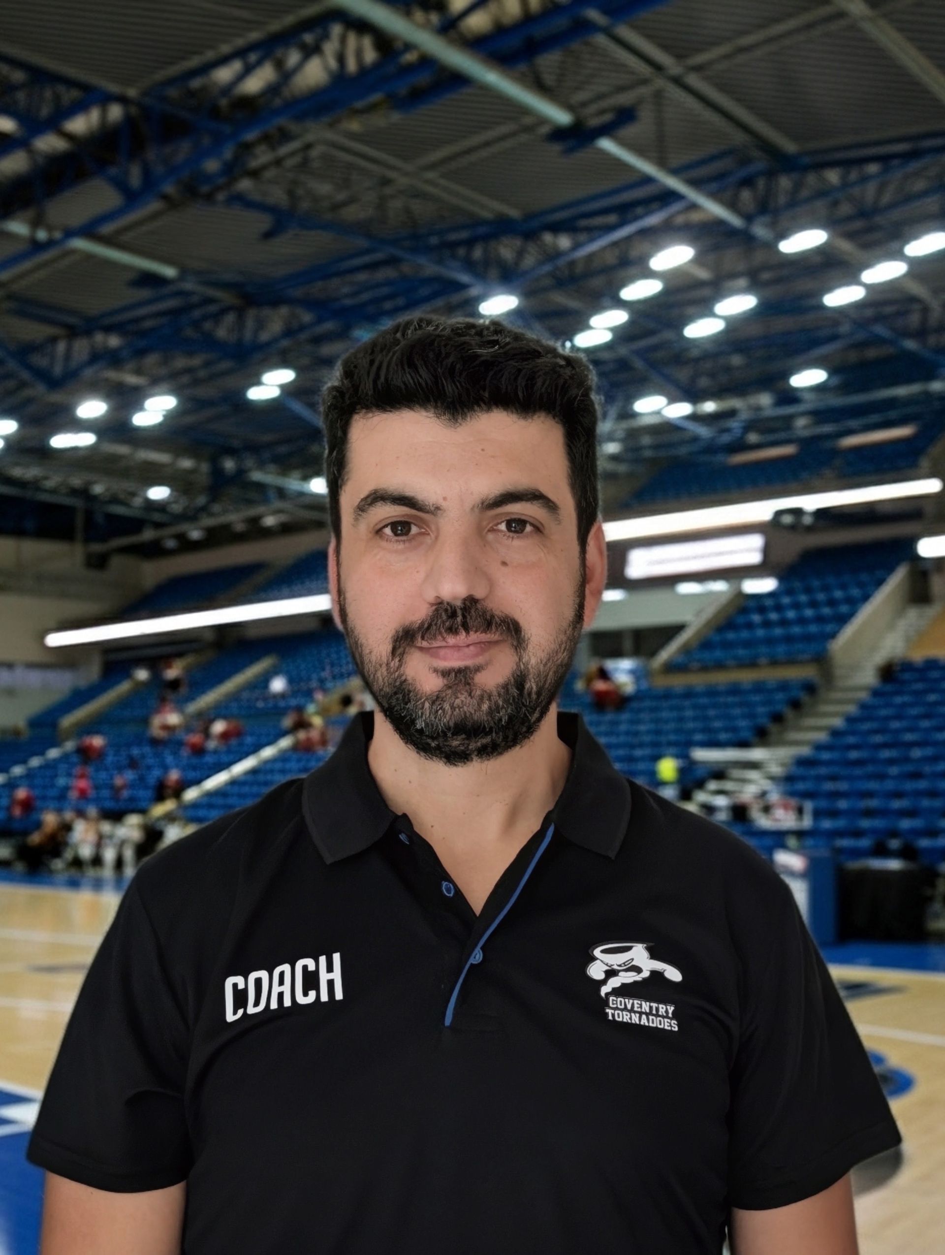 Man standing in a blue indoor arena, wearing a black coach polo shirt with a slight smile.