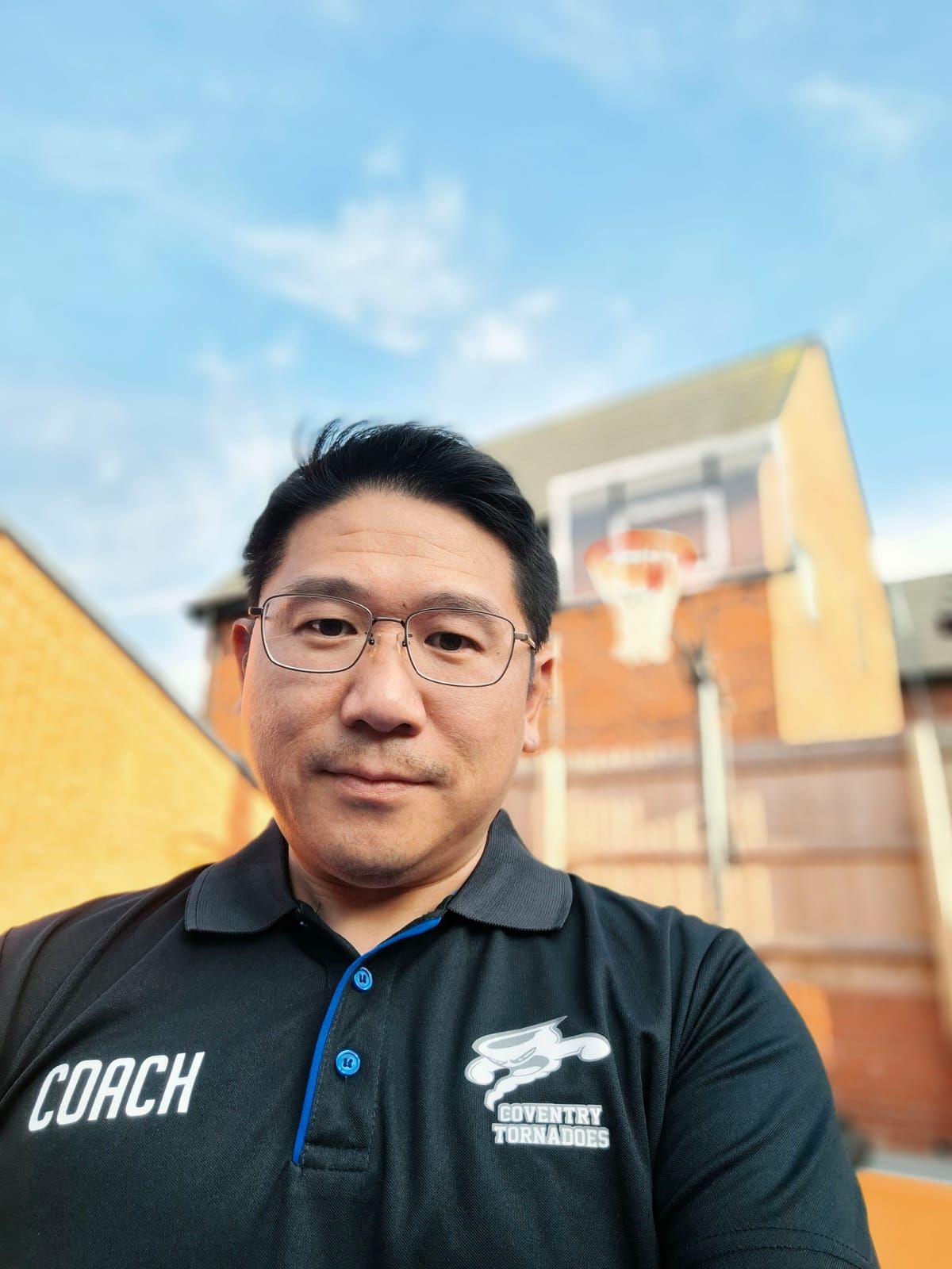 Man in black COACH shirt taking a selfie on an outdoor basketball court with hoop and blue sky