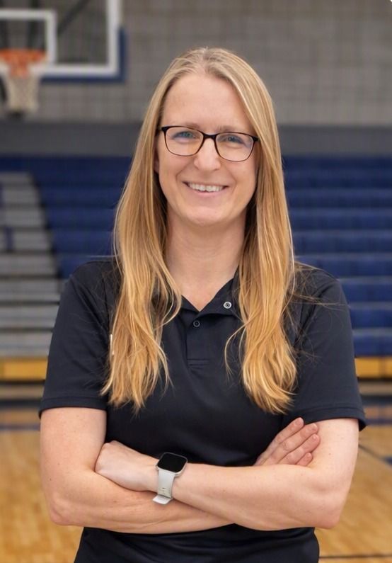 Smiling person with long blonde hair and glasses standing in a gym with arms crossed