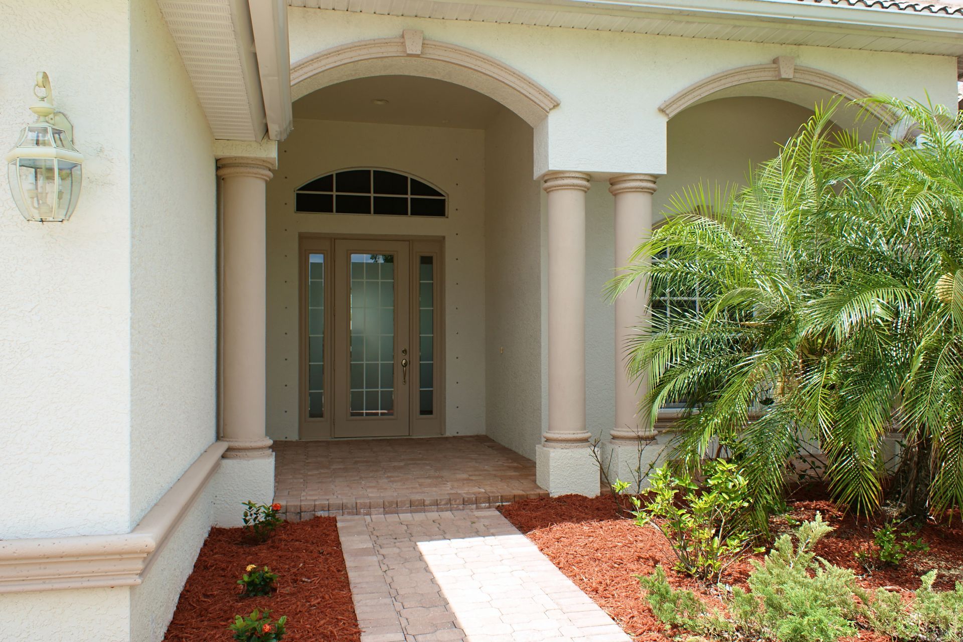 Tan house entrance with arched doorway, columns, and walkway lined with red mulch.