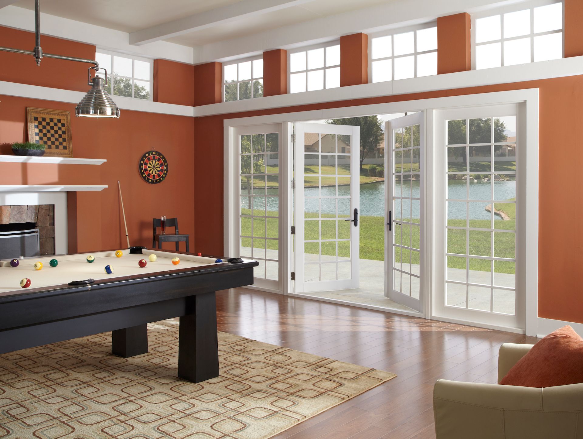 A game room with a pool table, orange walls, and large white-framed glass doors overlooking a lake.