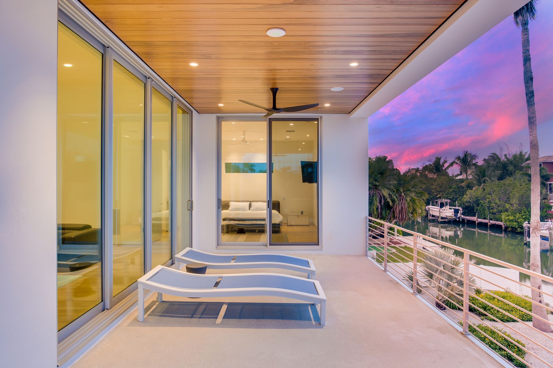 Balcony with two lounge chairs overlooking water at sunset, next to sliding glass doors.