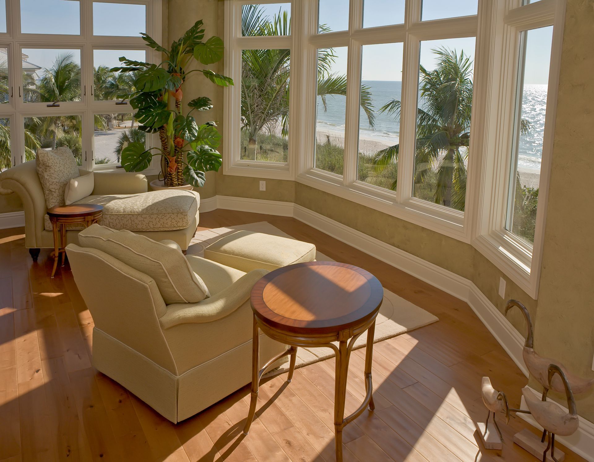 Bright sunroom with beach view, two armchairs, small tables, and a large leafy plant.