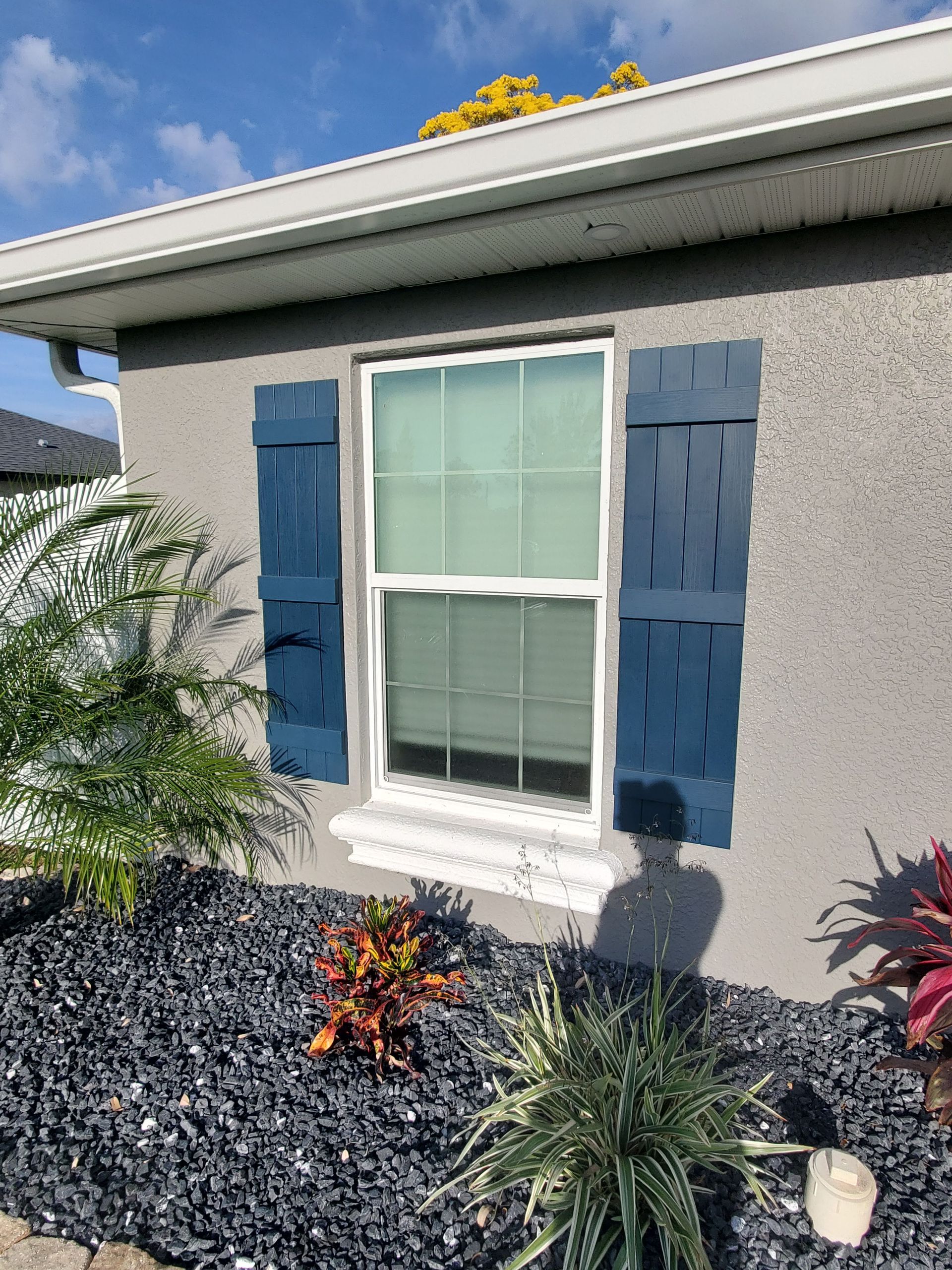 Blue shutters flank a window on a gray stucco house. Landscaping with dark rocks and plants is in front.