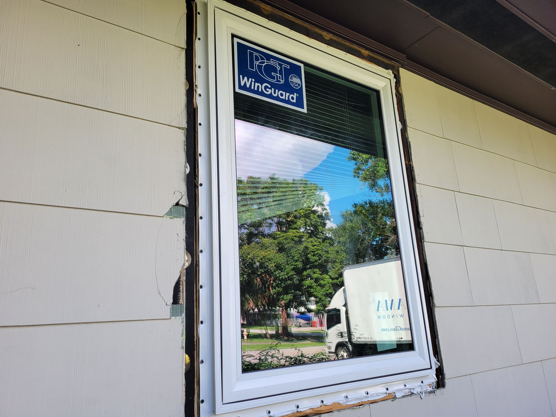 New white window installed on a house with white siding, reflecting trees and a truck.