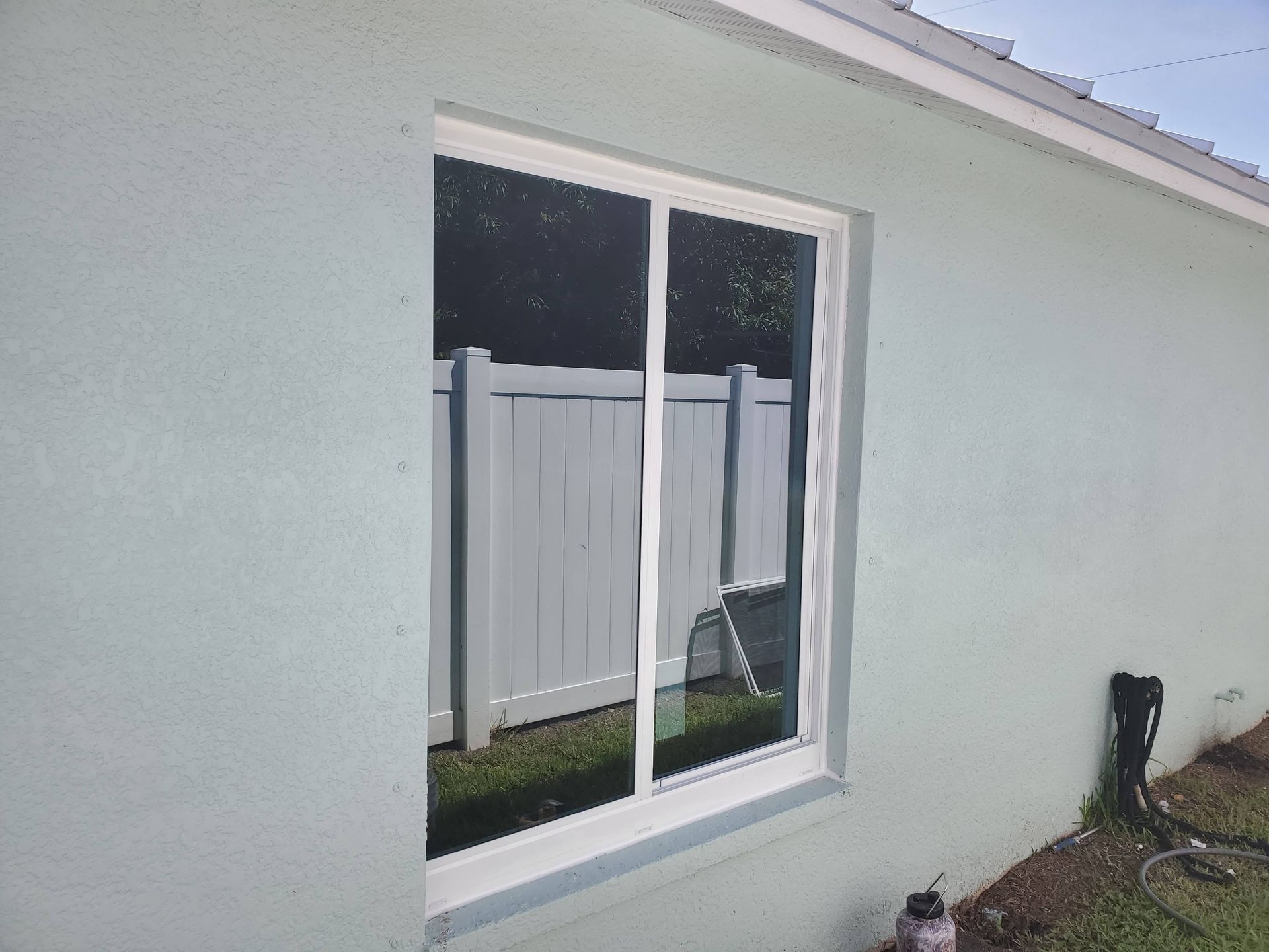 Sliding glass window on a pale blue stucco building, reflecting a white fence and grass.