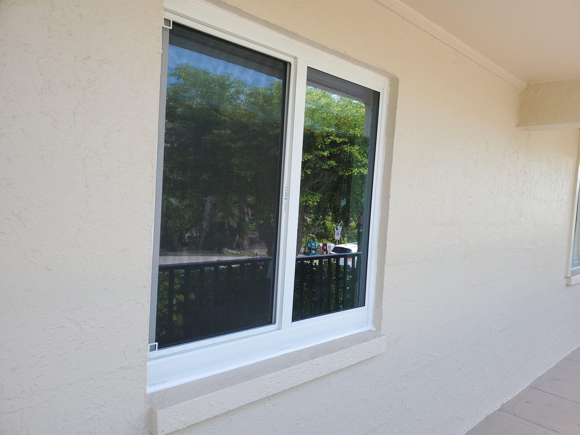 White-framed window with dark screen, reflecting a balcony and trees, set in a beige stucco wall.