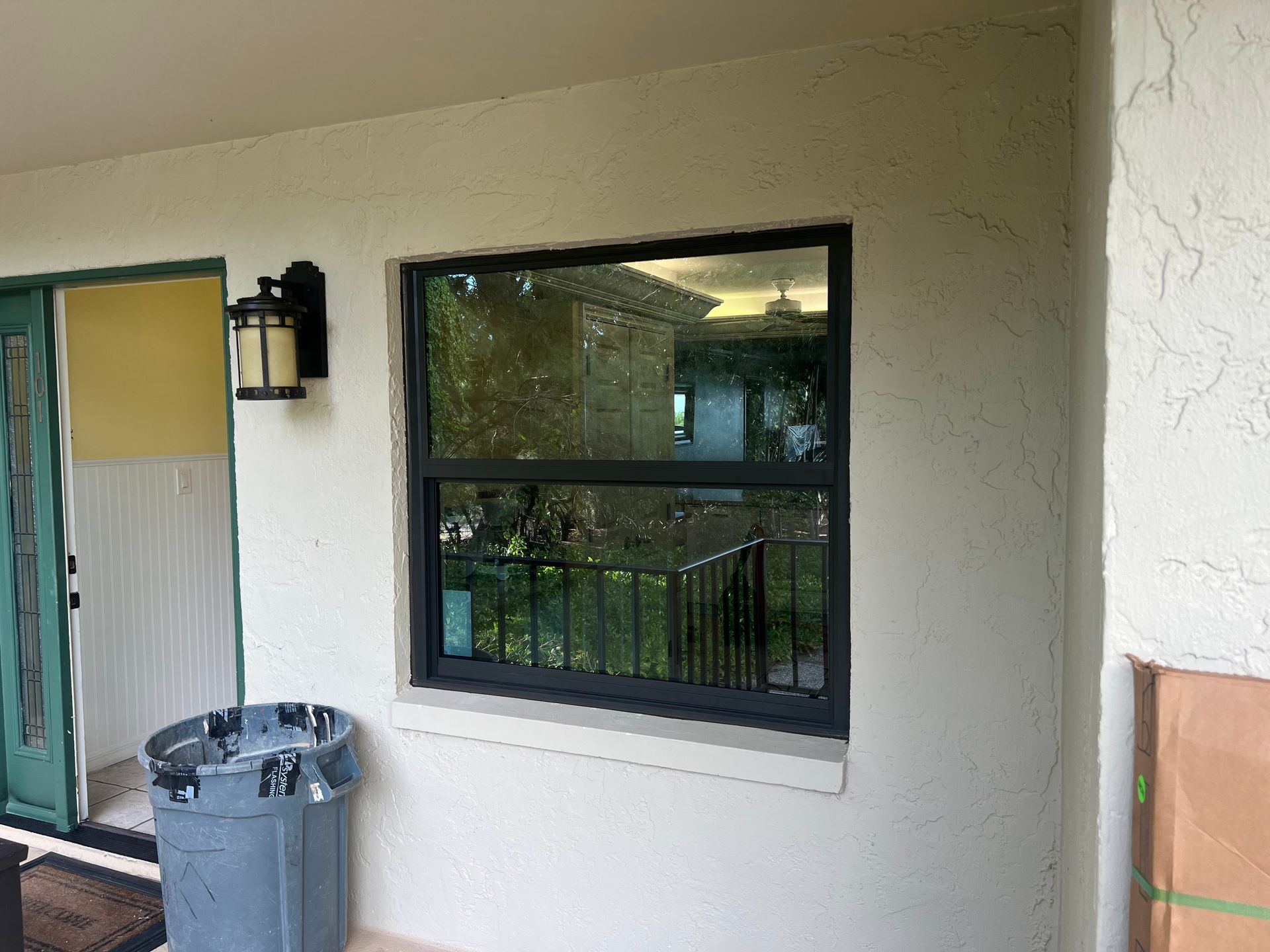Window with black frame on a beige wall, next to a trash can and a door, outdoor setting.
