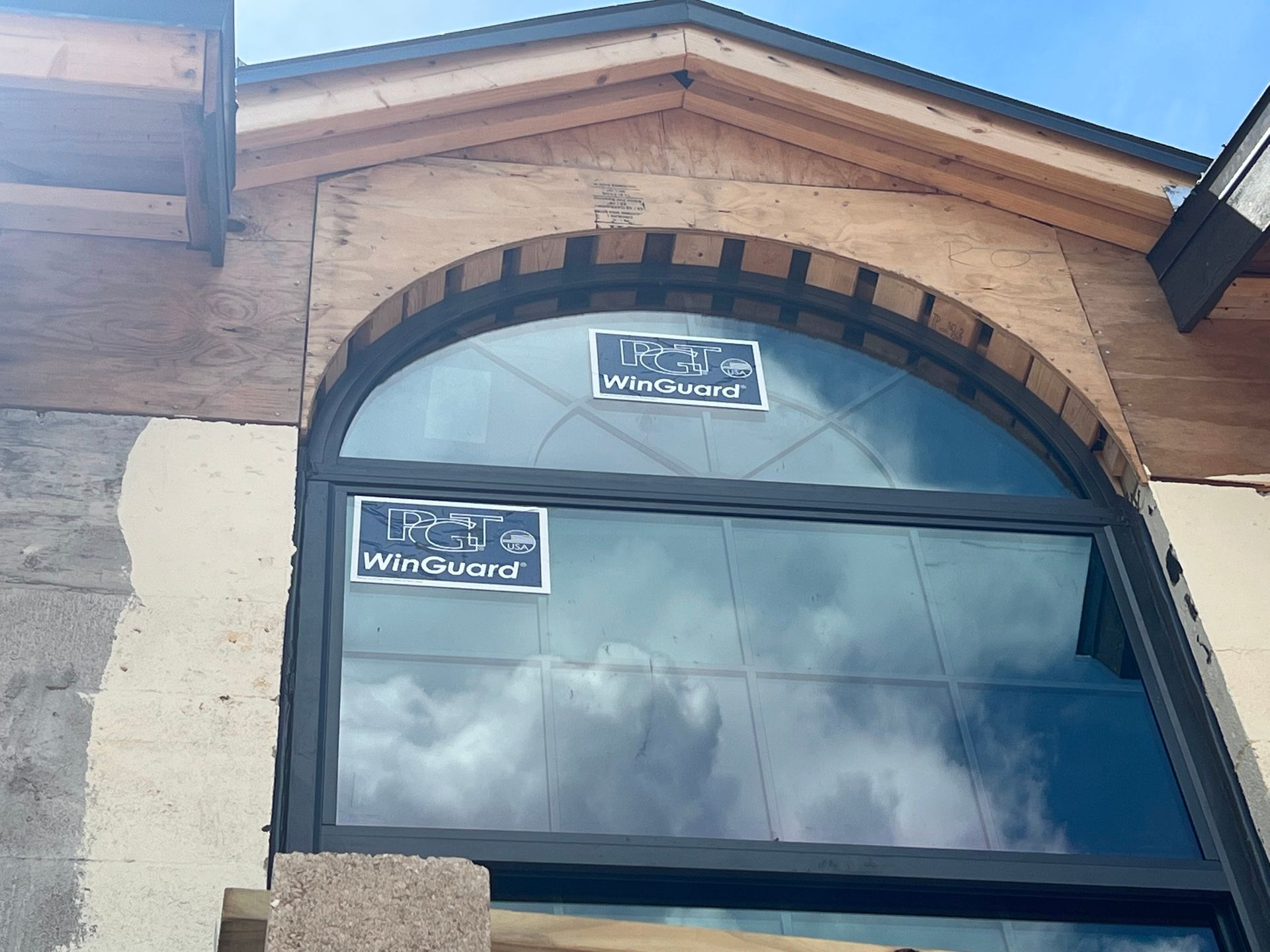Large, arched window installed in a building under construction. Dark frame, cloudy sky reflecting in the glass.