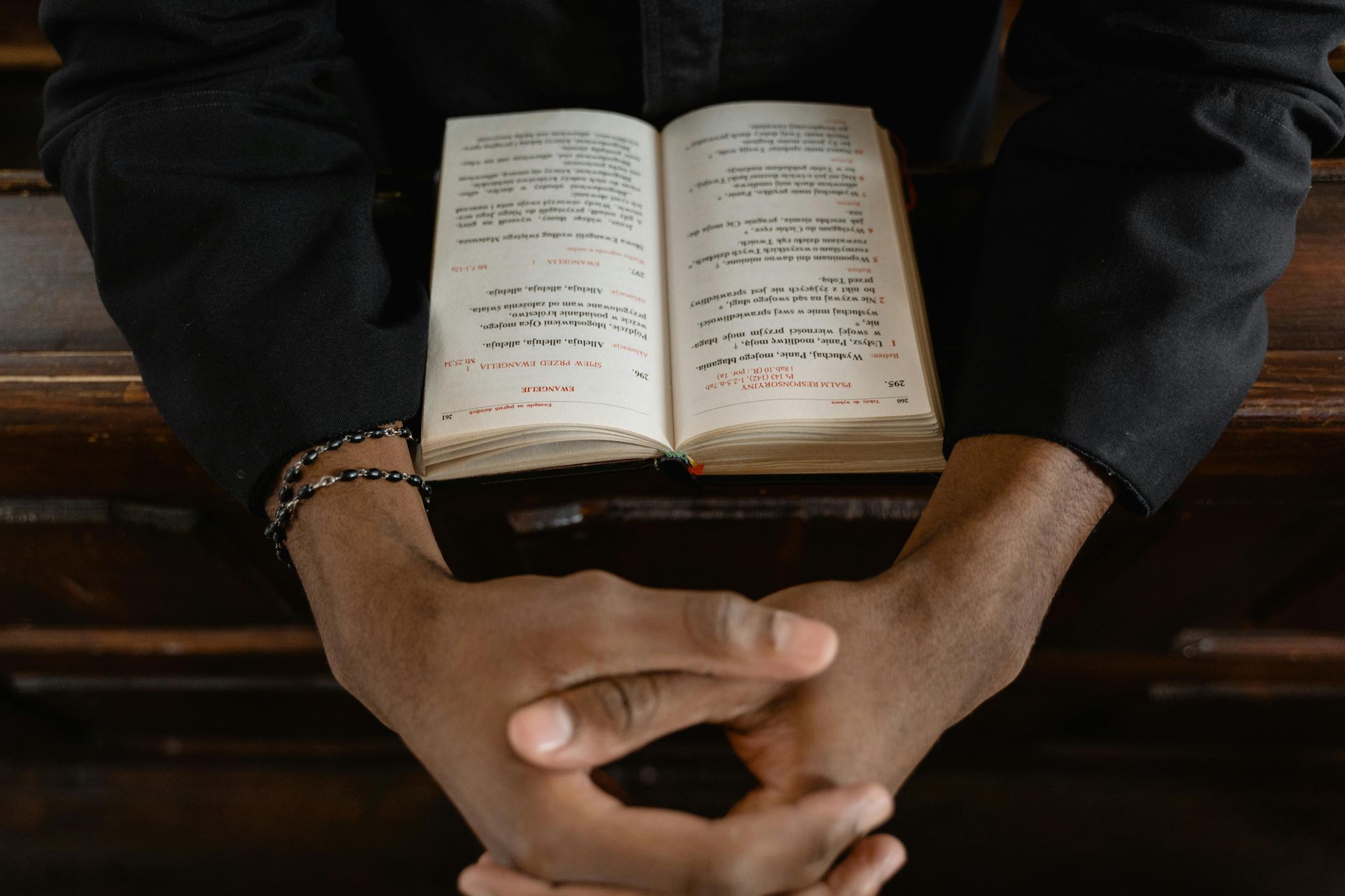 Hands clasped over an open book on a wooden surface; the person wears a black shirt.