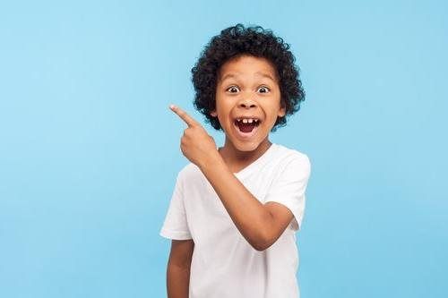 Boy with curly hair, in a white shirt, points and smiles with excitement. Blue background.