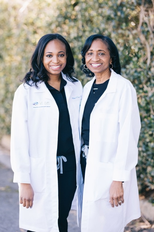 Two women in white lab coats smile, standing outside a building with green foliage in the background.