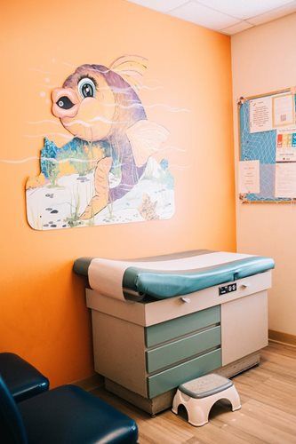 Pediatric exam room with fish mural, examination table, and footstool. Orange wall, blue and teal accents.
