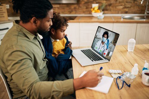 Man and child on a video call with a doctor on laptop. Man takes notes at a table.