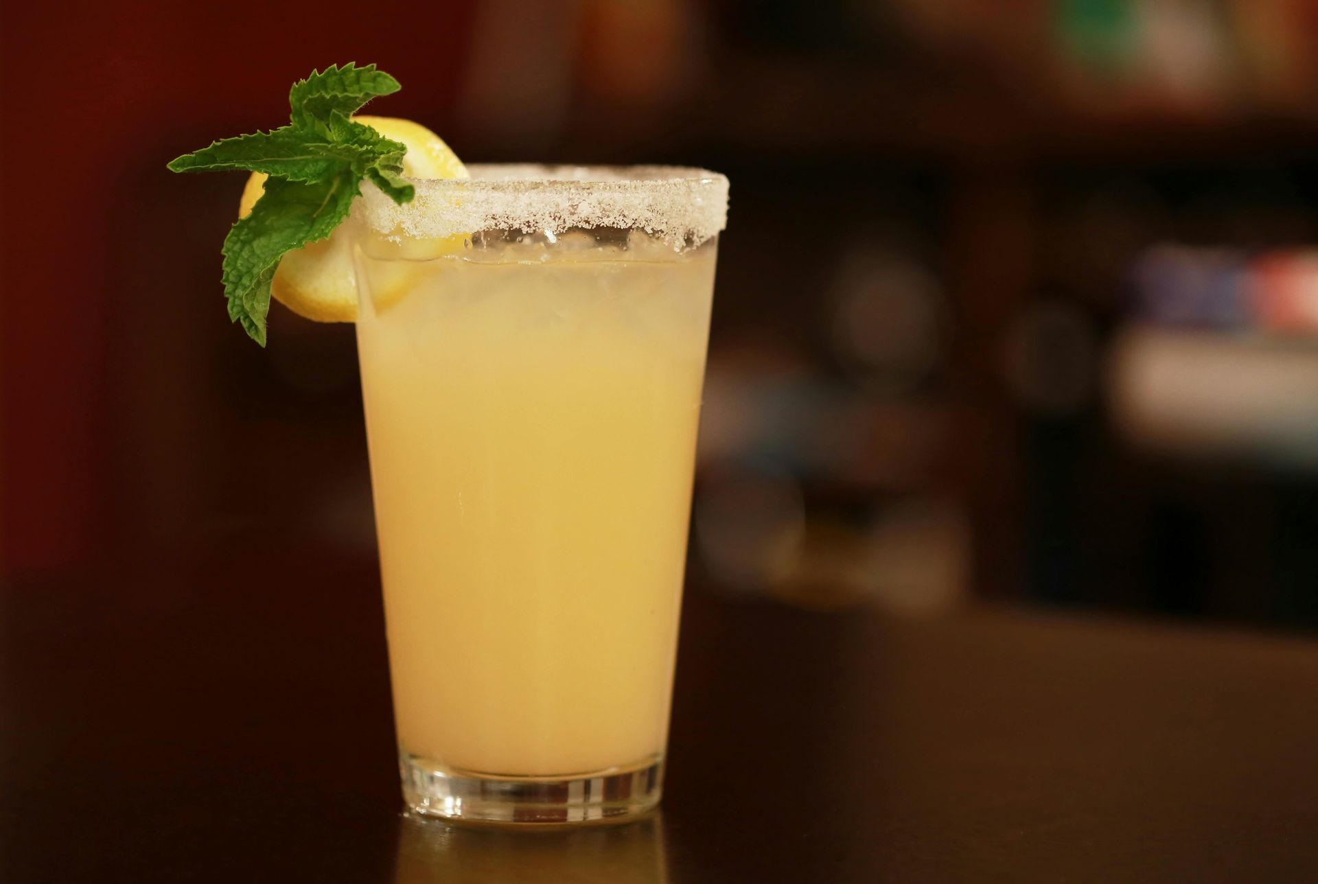 Tall glass with a yellow cocktail, sugar rim, lemon slice, and mint sprig, on a dark bar.