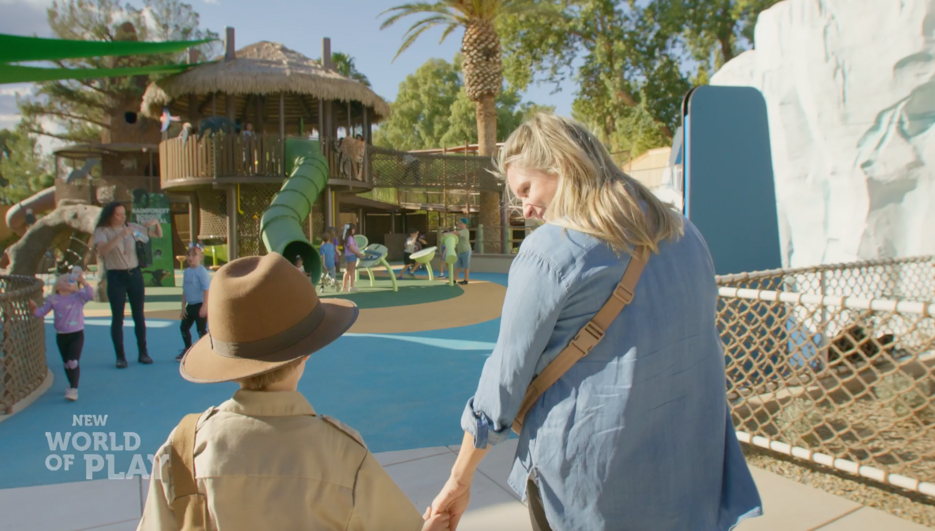 A woman is holding the hand of a child in a playground at Reid Park Zoo.