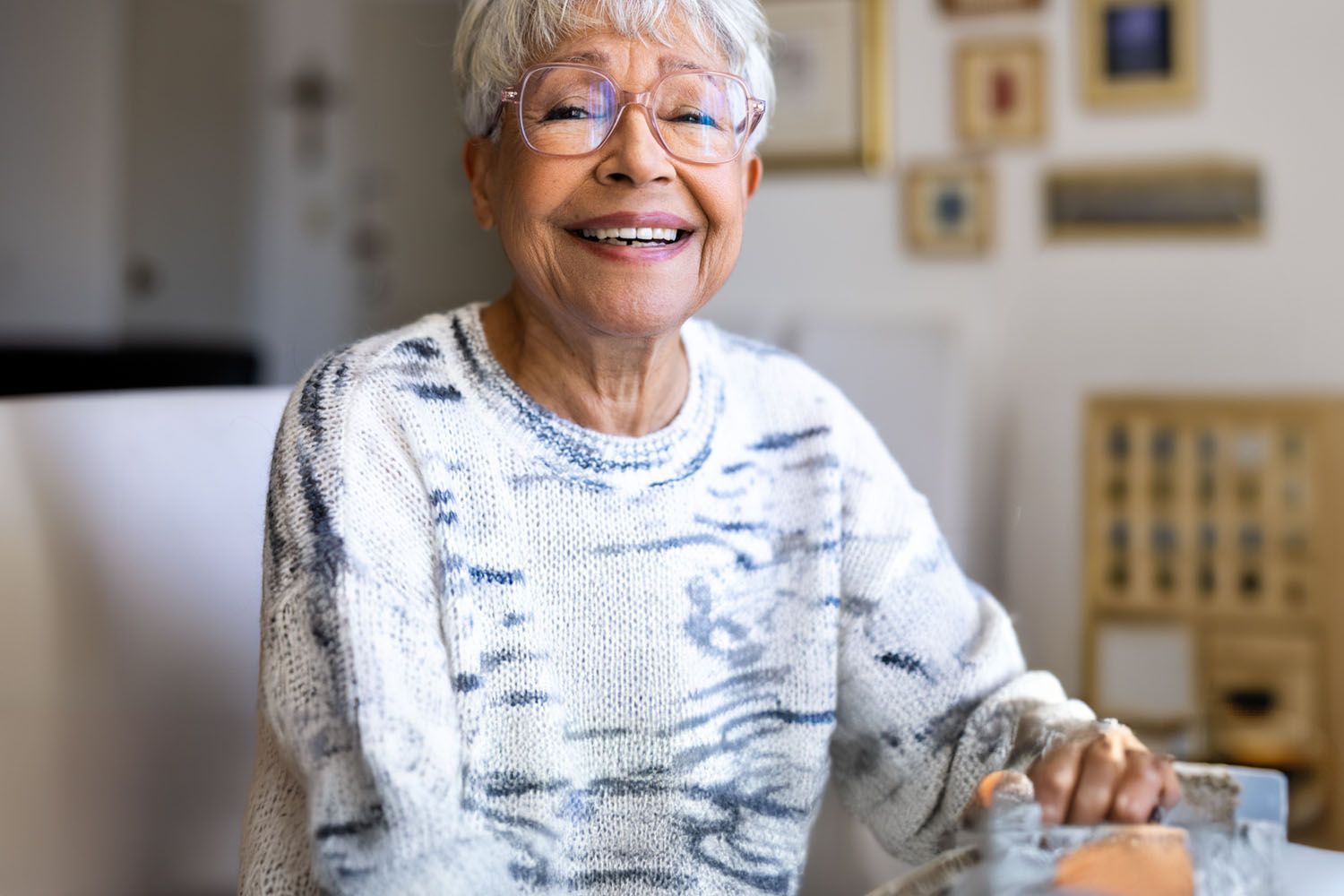 An elderly woman is smiling while sitting at a table with a glass of water.