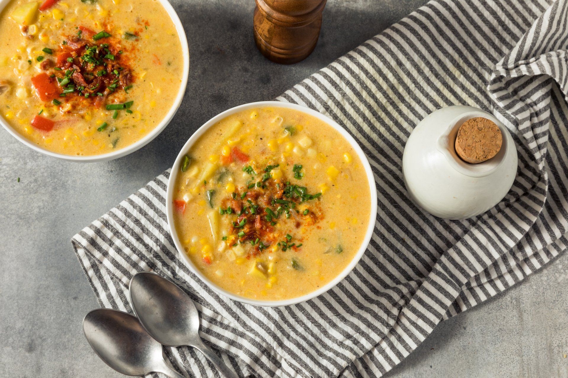 Two bowls of corn chowder with spoons on a striped towel on a table.