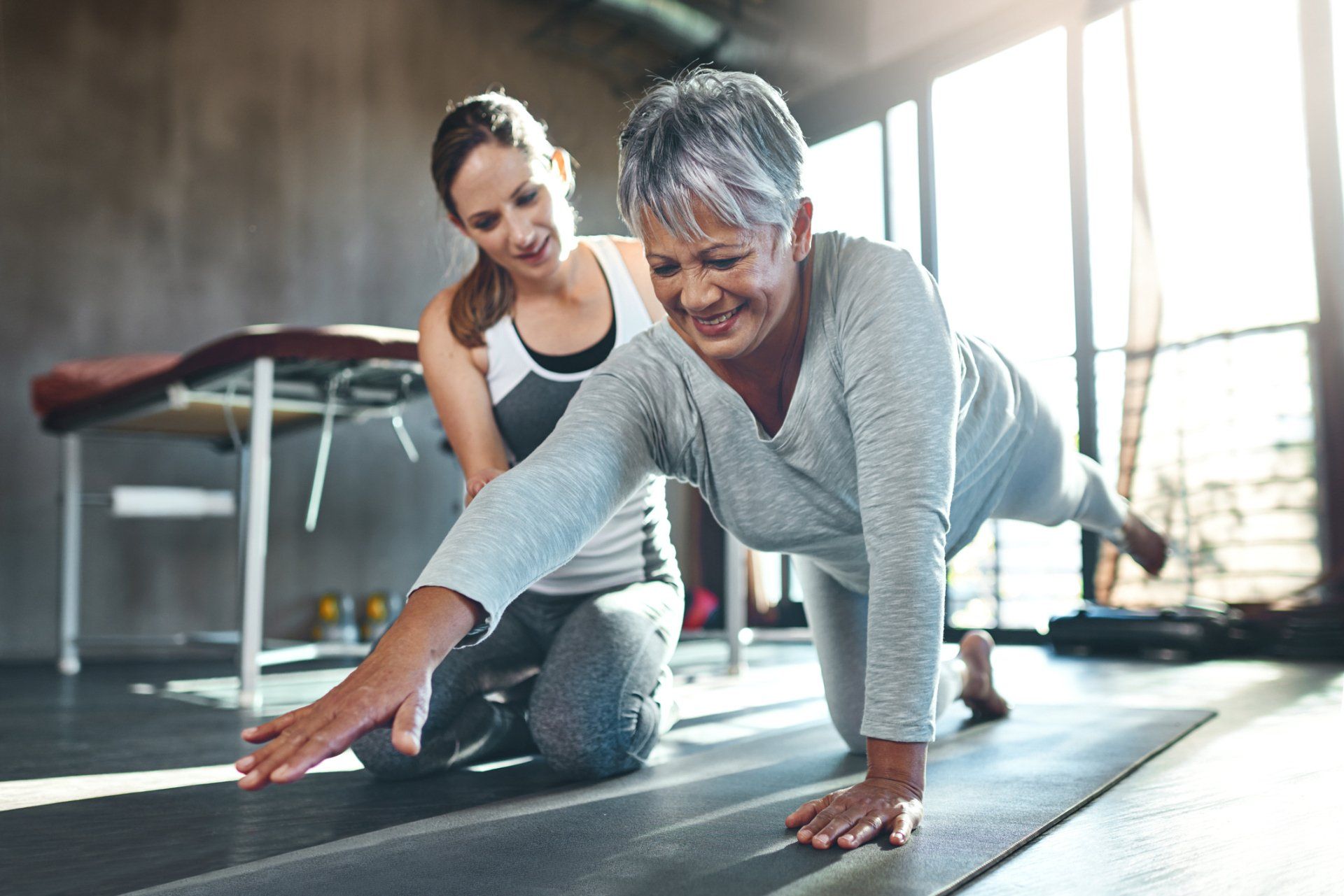 Woman doing plank exercise with a trainer in a gym, on a mat by large windows.