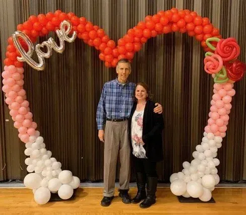 A man and a woman are standing in front of a heart shaped arch made of balloons.