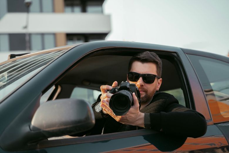 Man in sunglasses holding camera, leaning out car window, taking photo.