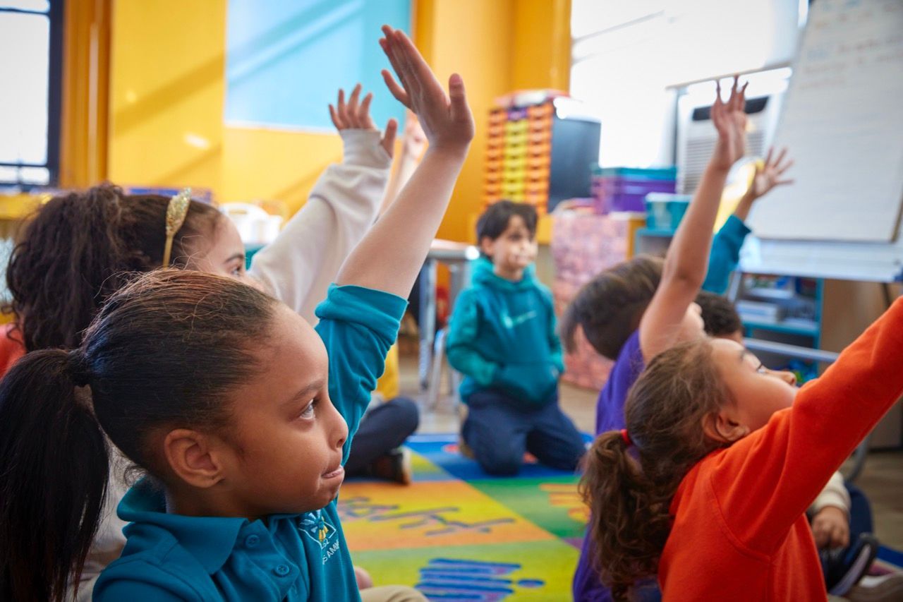 Children raising hands on a colorful classroom rug during group time