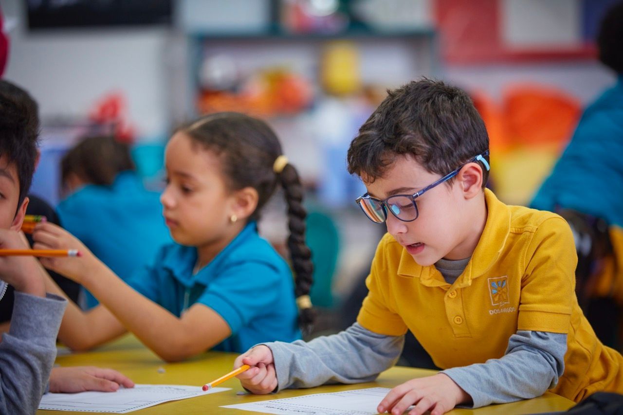 Children painting at a classroom table, including a boy in a yellow shirt and glasses drawing with a pencil.