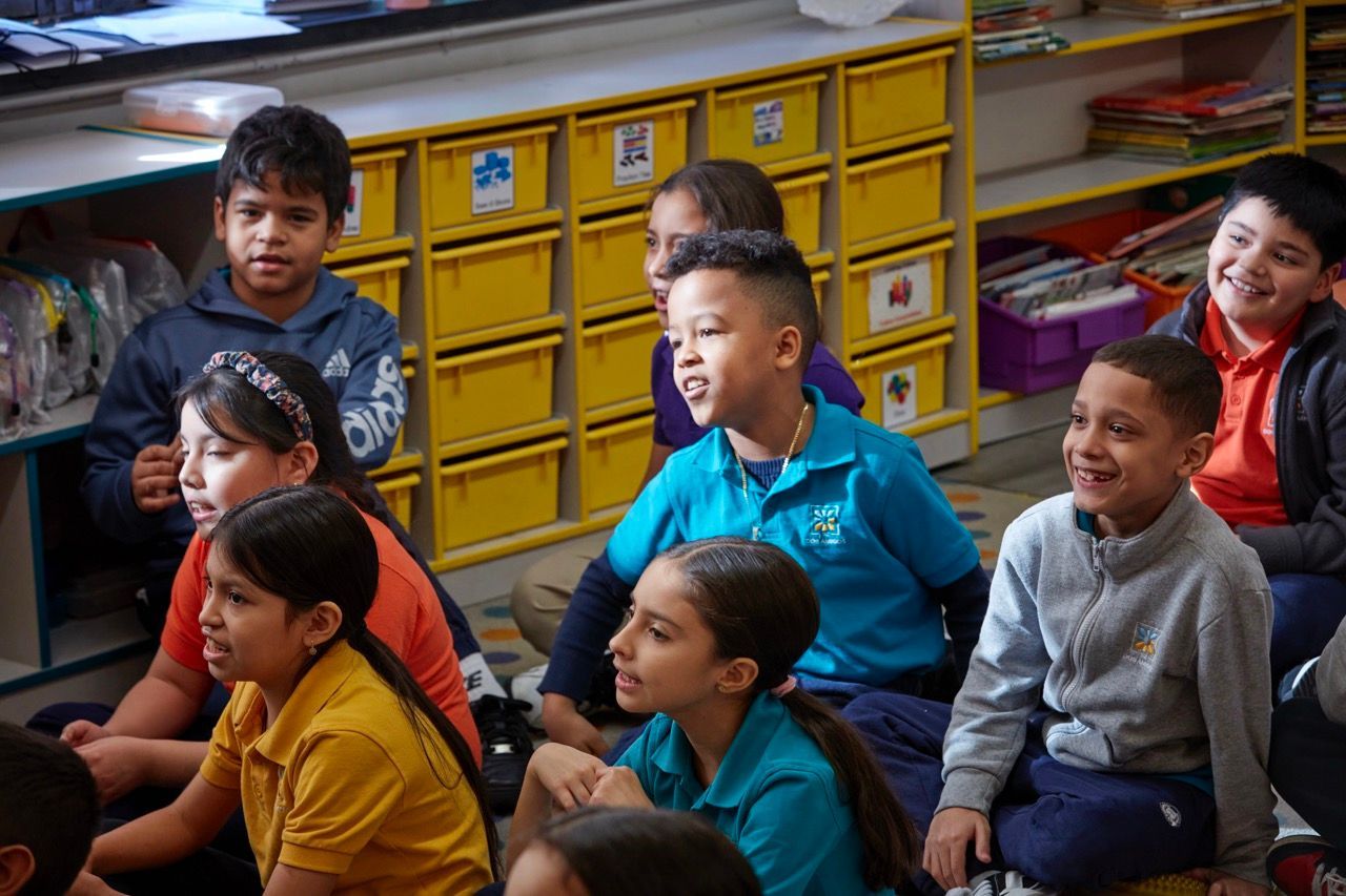 Children sitting together in a classroom, with yellow storage drawers in the background.