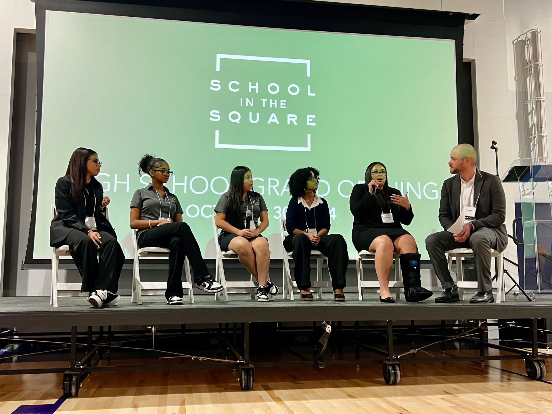 Panel discussion on stage with six seated speakers and a school logo backdrop