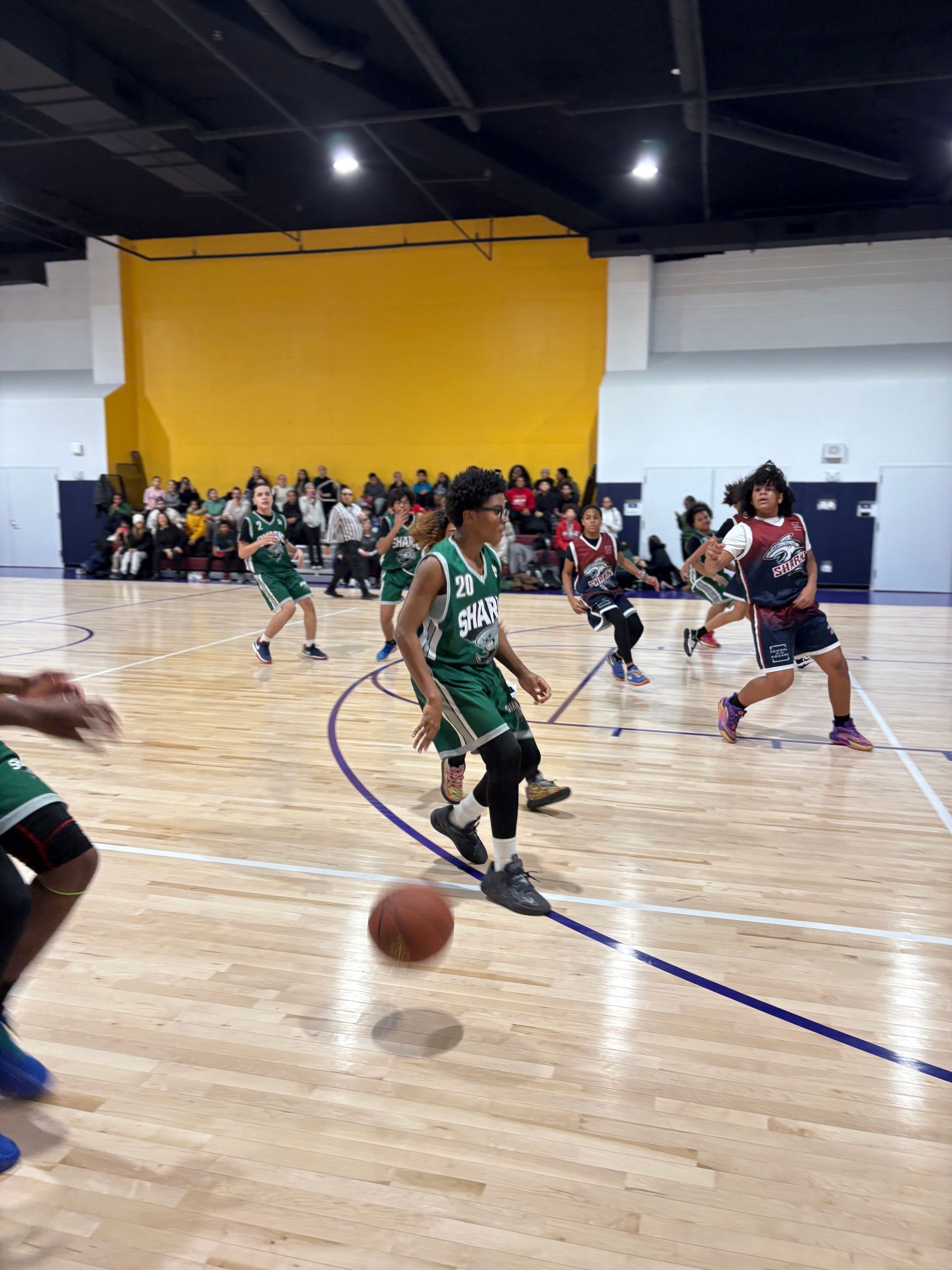 Basketball game on an indoor court, with players dribbling near the sideline and spectators watching nearby.