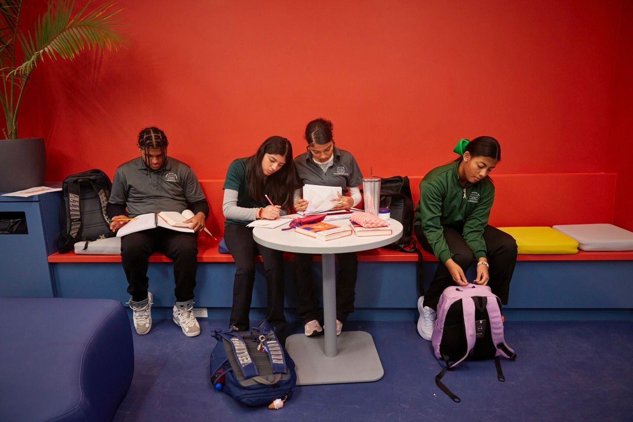 People sitting at a white table in a red-walled room, looking down at papers and a pink backpack nearby