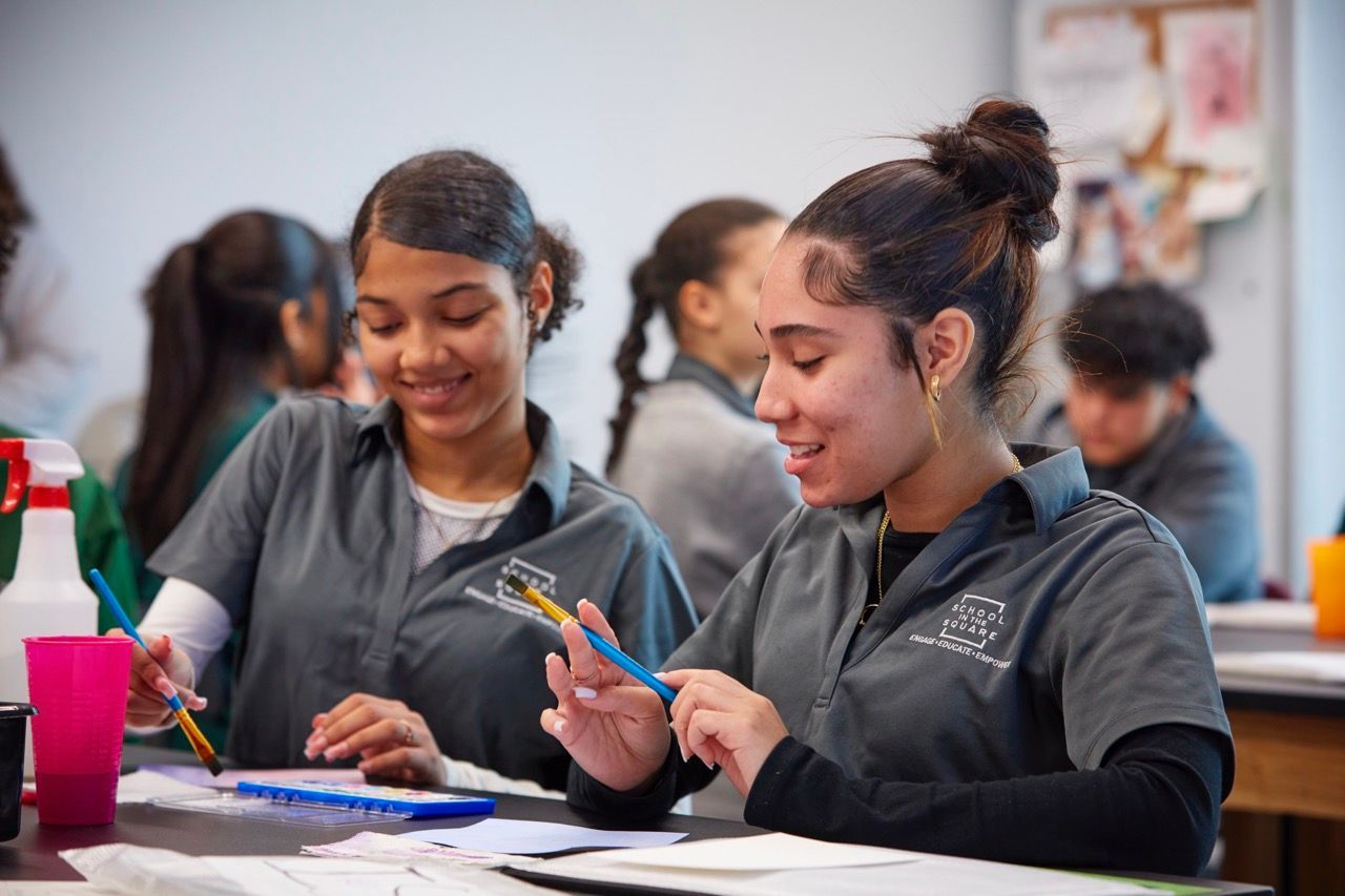 Students working together at a classroom desk, smiling and writing with colored pencils