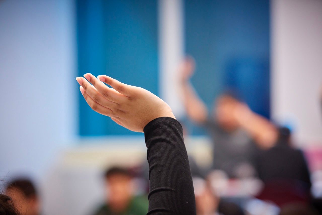 Raised hand in a classroom or meeting, with blurred people and blue walls in the background