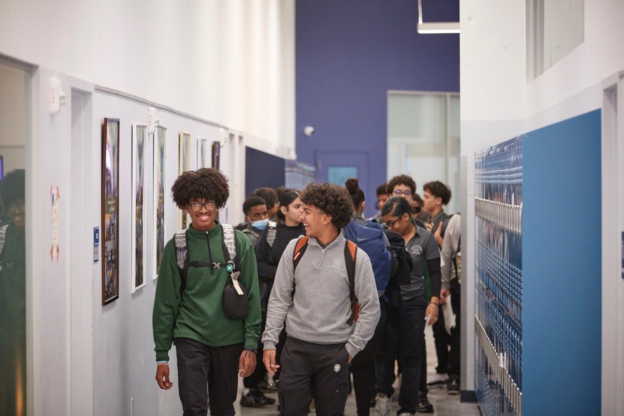 Students walking in a school hallway past blue lockers and white walls