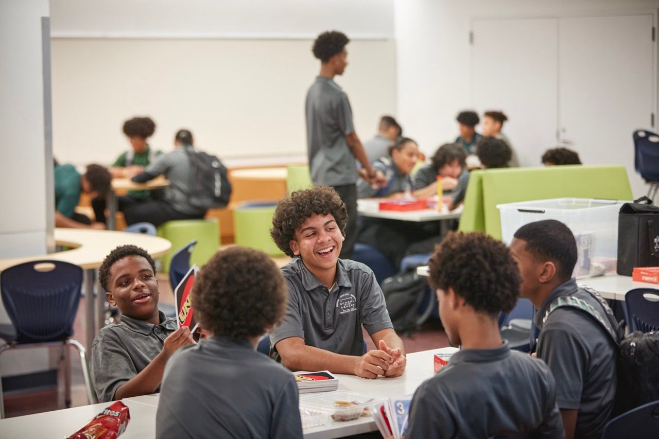 Students chatting at cafeteria tables in a bright dining hall, with trays and green tablecloths nearby.