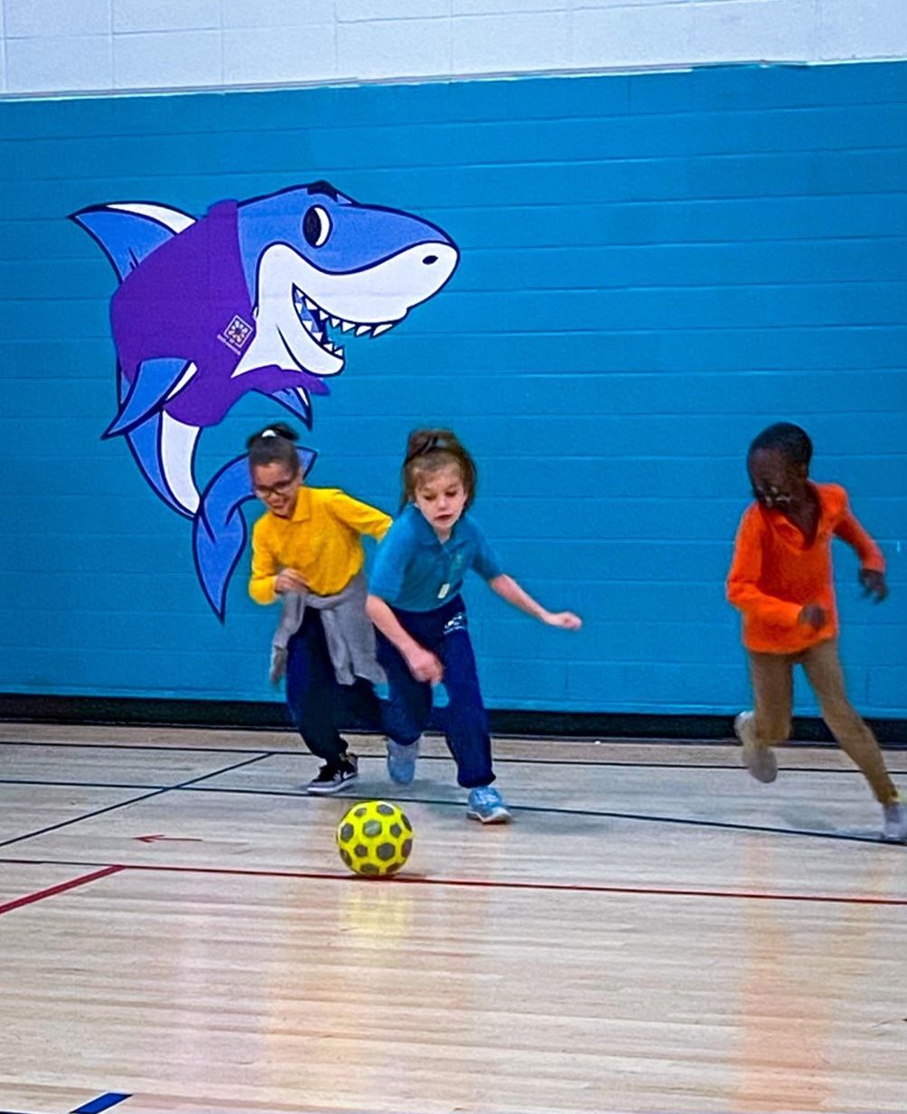 Children playing indoor soccer in a gym, with a yellow ball and a shark mural on the blue wall